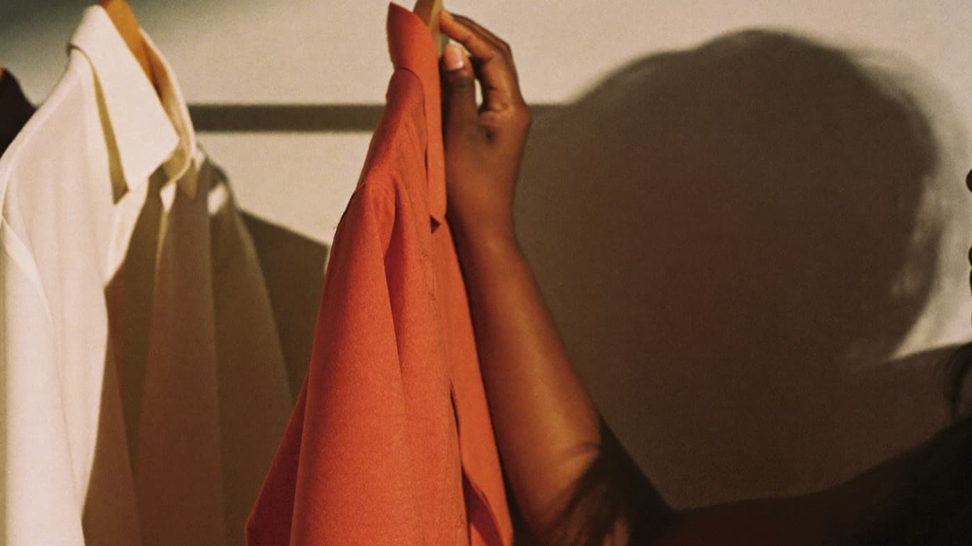 A hand lifting a coral silk shirt from a brass rail in hard afternoon light, cream and neutral garments hanging alongside