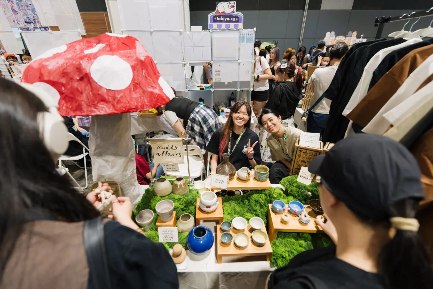 Two sellers smile behind a table of handmade ceramic cups and vases set on moss, beside a large paper mushroom, at Artsobi 3.0 in Suntec Singapore, August 2026