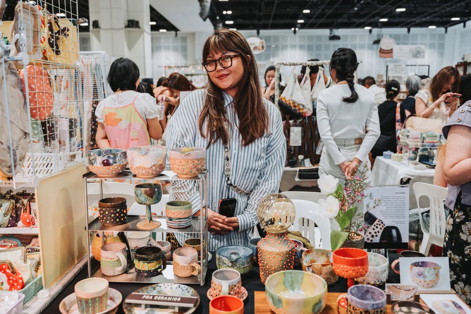 A ceramicist stands behind her stall of handmade cups, bowls and vases while shoppers browse the aisles at Asia Art & Craft Fair 2026 in Singapore