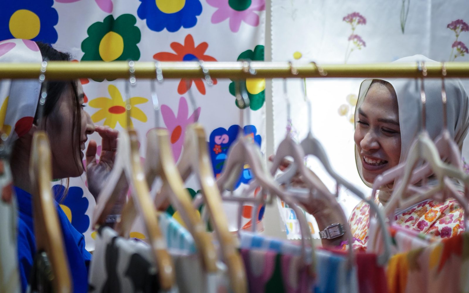 Two women laugh across a rail of clothes on hangers at a fashion booth at a past Celebfest in Singapore
