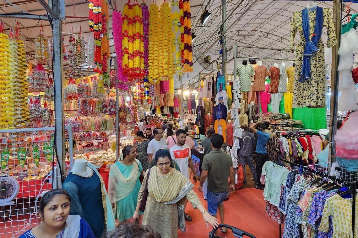 Shoppers walking a busy aisle inside the Deepavali bazaar tent in Little India, Singapore, lined with marigold garlands and Indian clothing stalls