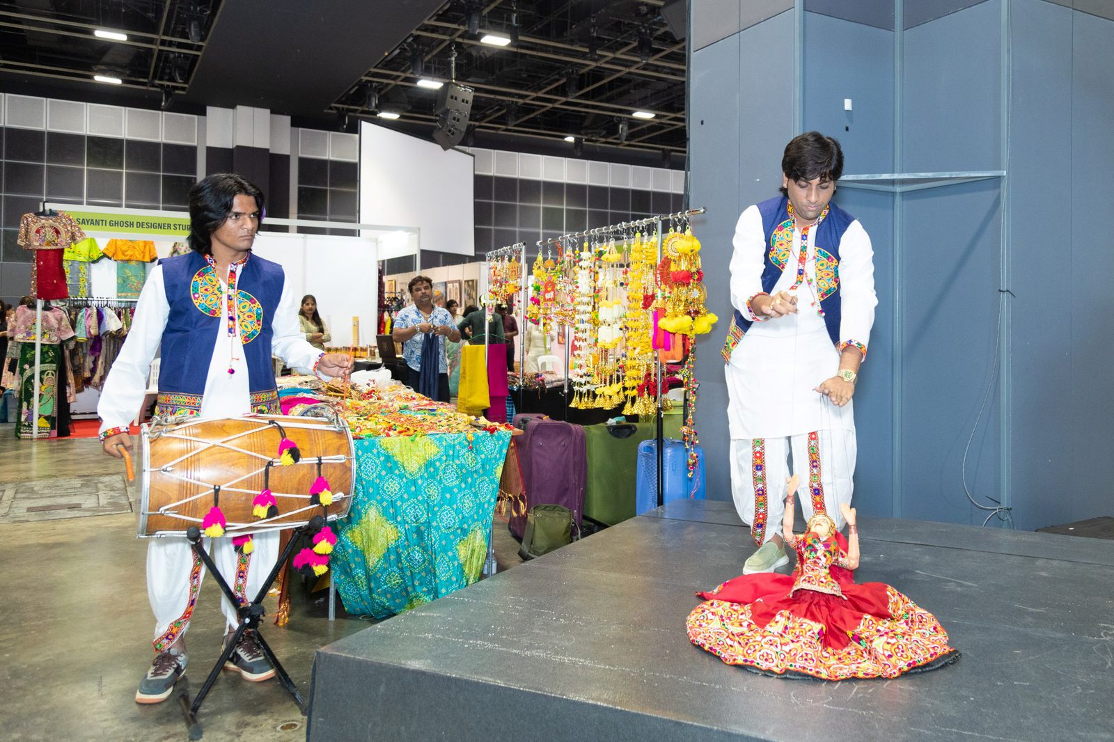 A puppeteer and a drummer in embroidered waistcoats perform beside stalls of garlands and clothing at Namaste Bharat in Suntec Singapore, October 2024