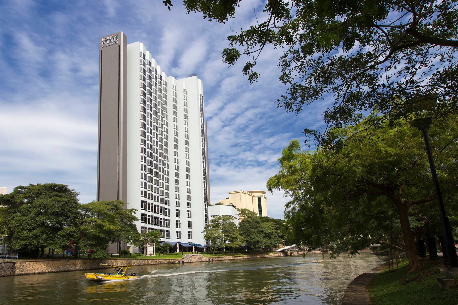 The Four Points by Sheraton Singapore, Riverview hotel tower seen across the Singapore River by day, with a river boat passing its tree-lined bank