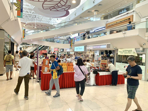 Red-skirted vendor tables in the Thomson Plaza atrium with shoppers browsing, Singapore