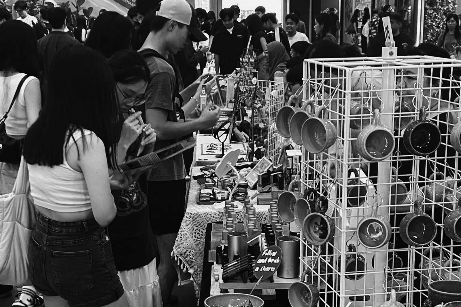 Black-and-white photograph of shoppers crowding a row of stalls with handmade pottery mugs and accessories at The Hammock Market in the Aperia Mall atrium, Singapore