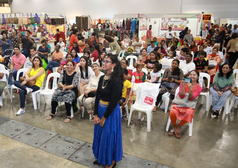 Seated audience watches a presenter in a blue skirt beside market stalls at Zak Salaam India Expo, Singapore Expo